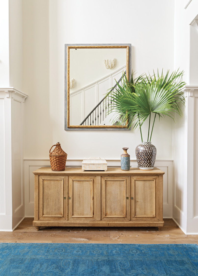 AWASH IN WHITE: In the foyer, a sideboard from Acquisitions Interiors mimics the wainscotting design. For subtle contrast and depth, Allison painted the walls Benjamin Moore’s “Swiss Coffee” and the trim “Simply White.” A mirror from Alexandra AD reflects the grand staircase.