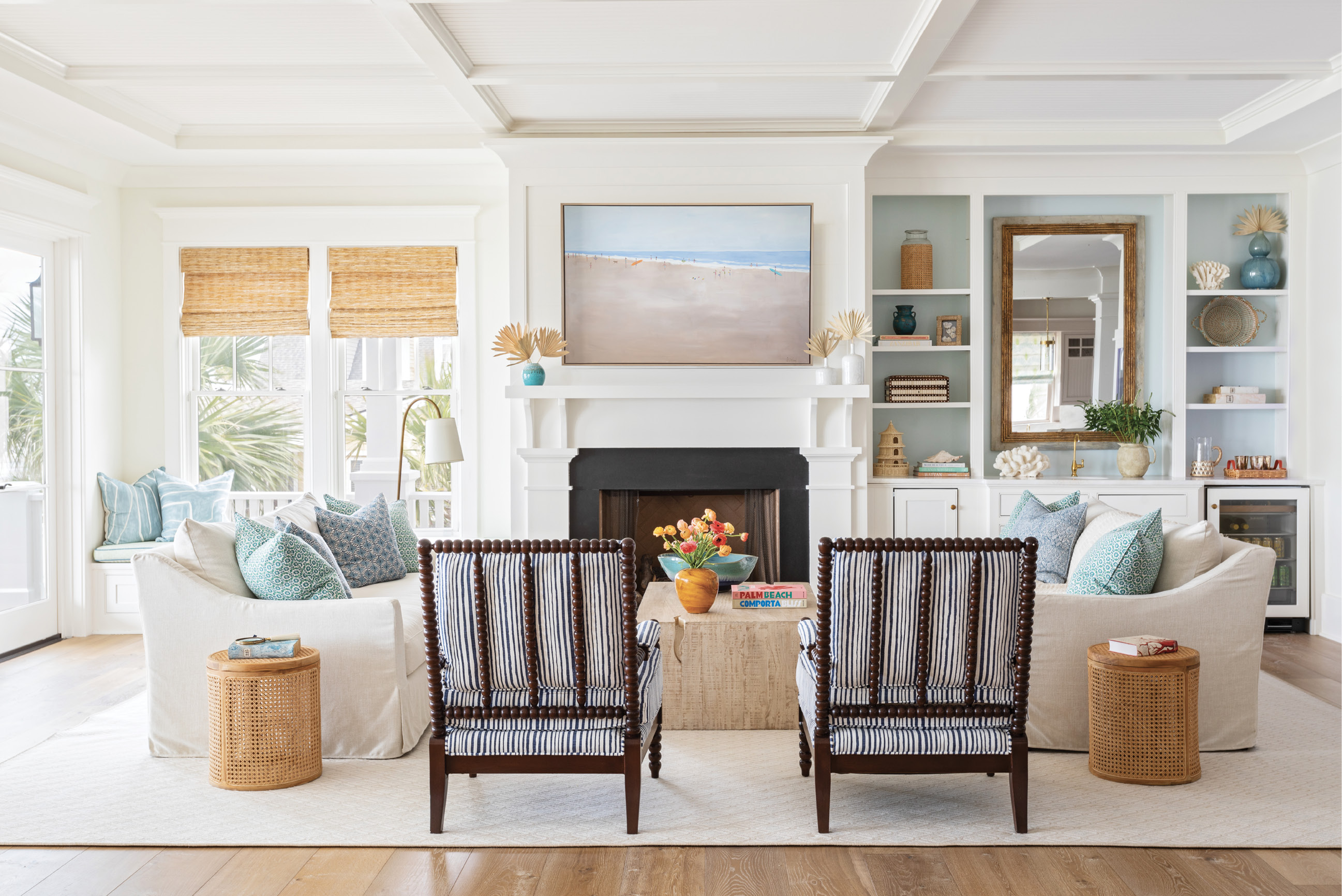 OCEANSIDE OASIS: In the heart of the home, natural elements—like a wooden coffee table from Celadon, cane end tables from GDC Home, and a wool rug—mix with performance fabric-outfitted furnishings for an unfussy, beach-friendly vibe. A beach scene by Shannon Wood, pillows from Celadon, and the wet bar’s back wall—painted Benjamin Moore’s “Ocean Air”—underscore the sea-inspired palette.