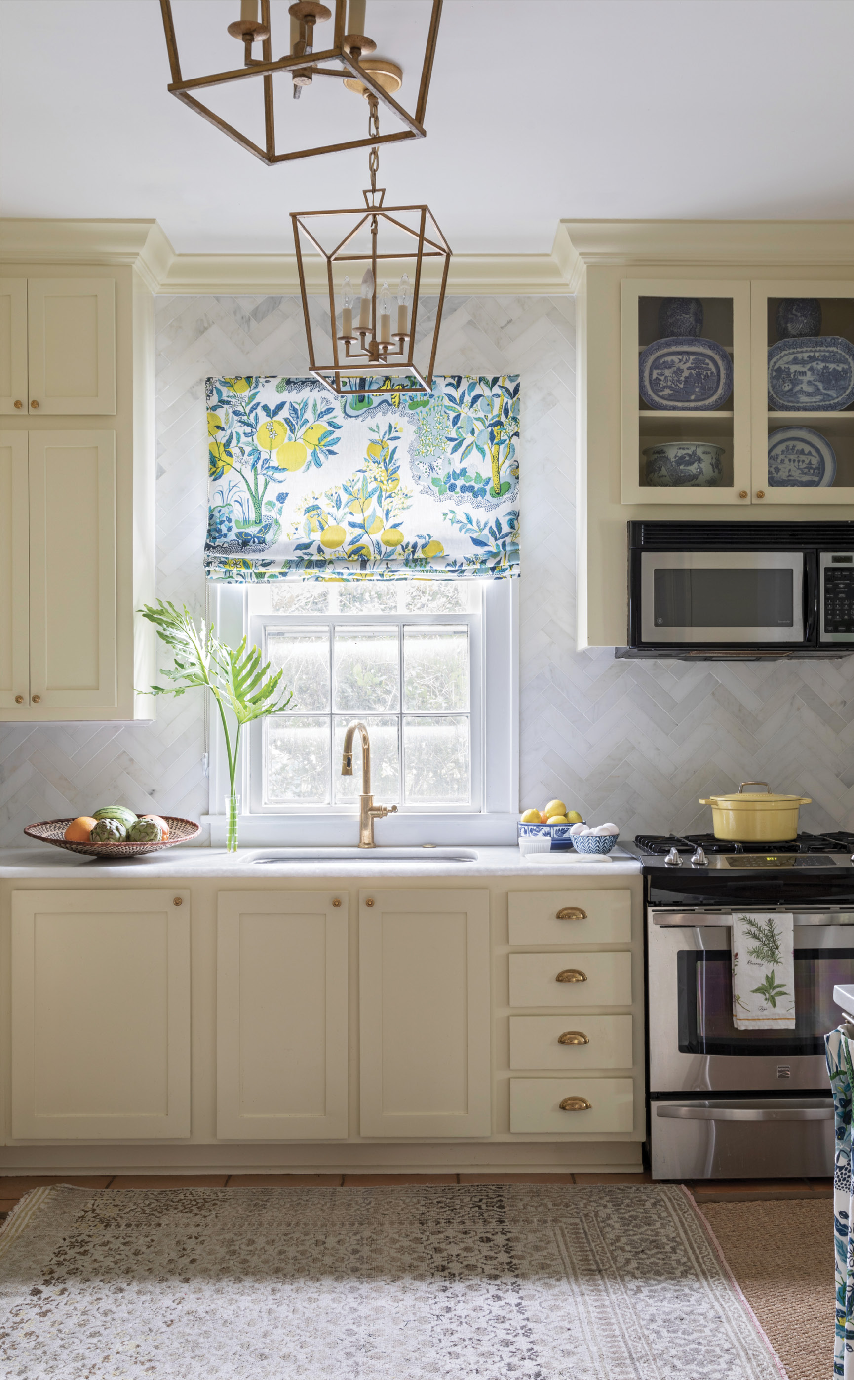 Bright Ideas: A colorful Schumacher fabric, hanging above the window and concealing a washer and dryer (above), makes a bold statement in this traditional cottage kitchen. Soft yellow Farrow &amp; Ball “Pale Hound” paint paired with brass hardware on the cabinets creates a vintage feel, while a white marble backsplash in a herringbone pattern and a giclée print by Hayley Mitchell add touches of modern luxury.