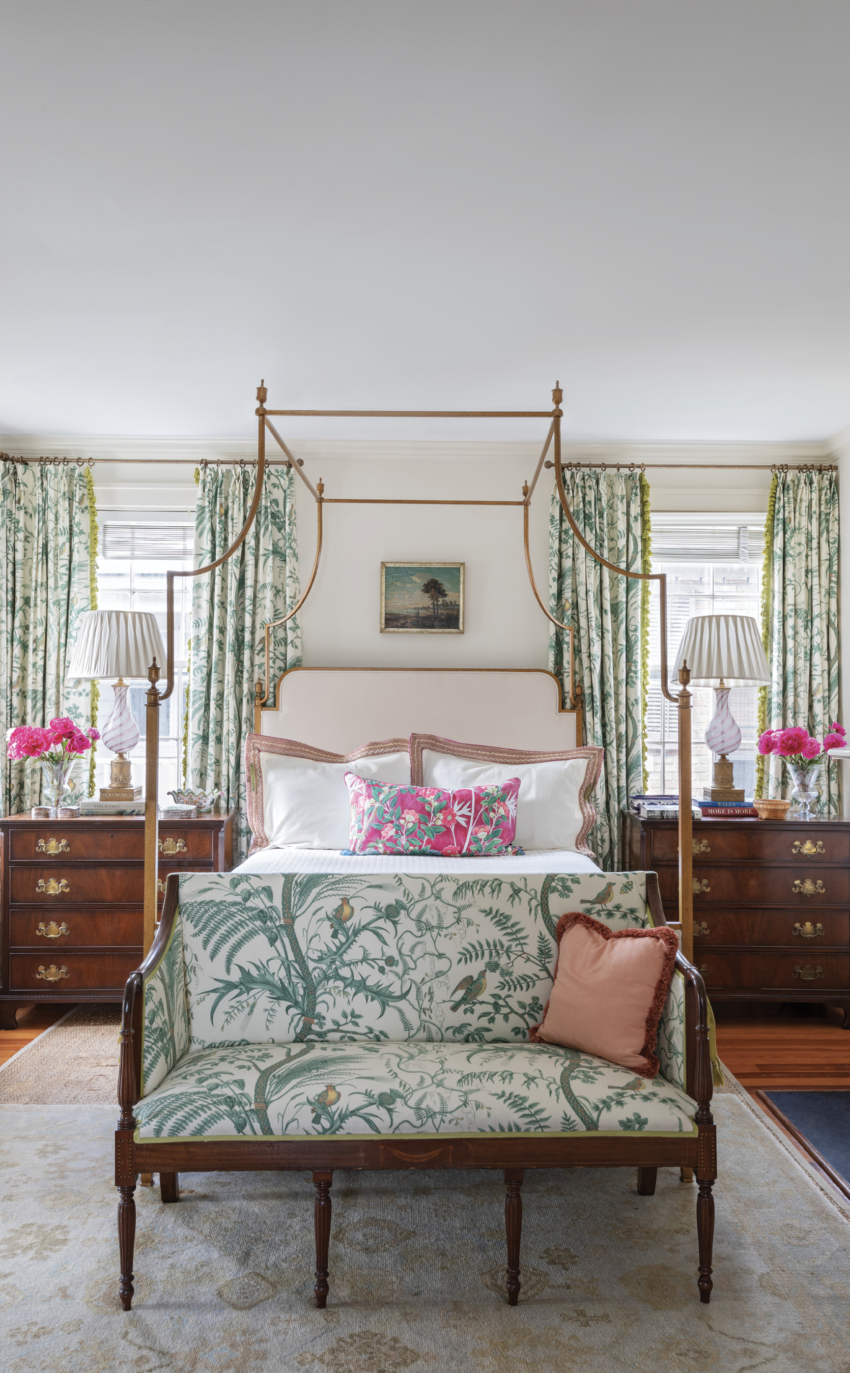 FIT FOR A QUEEN: A quieter color palette in the primary bedroom creates a soothing, luxurious space. The delicate design of the Brunschwig &amp; Fils “Bird &amp; Thistle” toile fabric for the curtains and the antique Sheraton settee, the brass four-poster bed, and vintage Murano glass lamps combine to produce a light and feminine feel. The theme continues in the bath with a white Carrara marble tiled tub and vanity highlighted with accents of Kelly green.