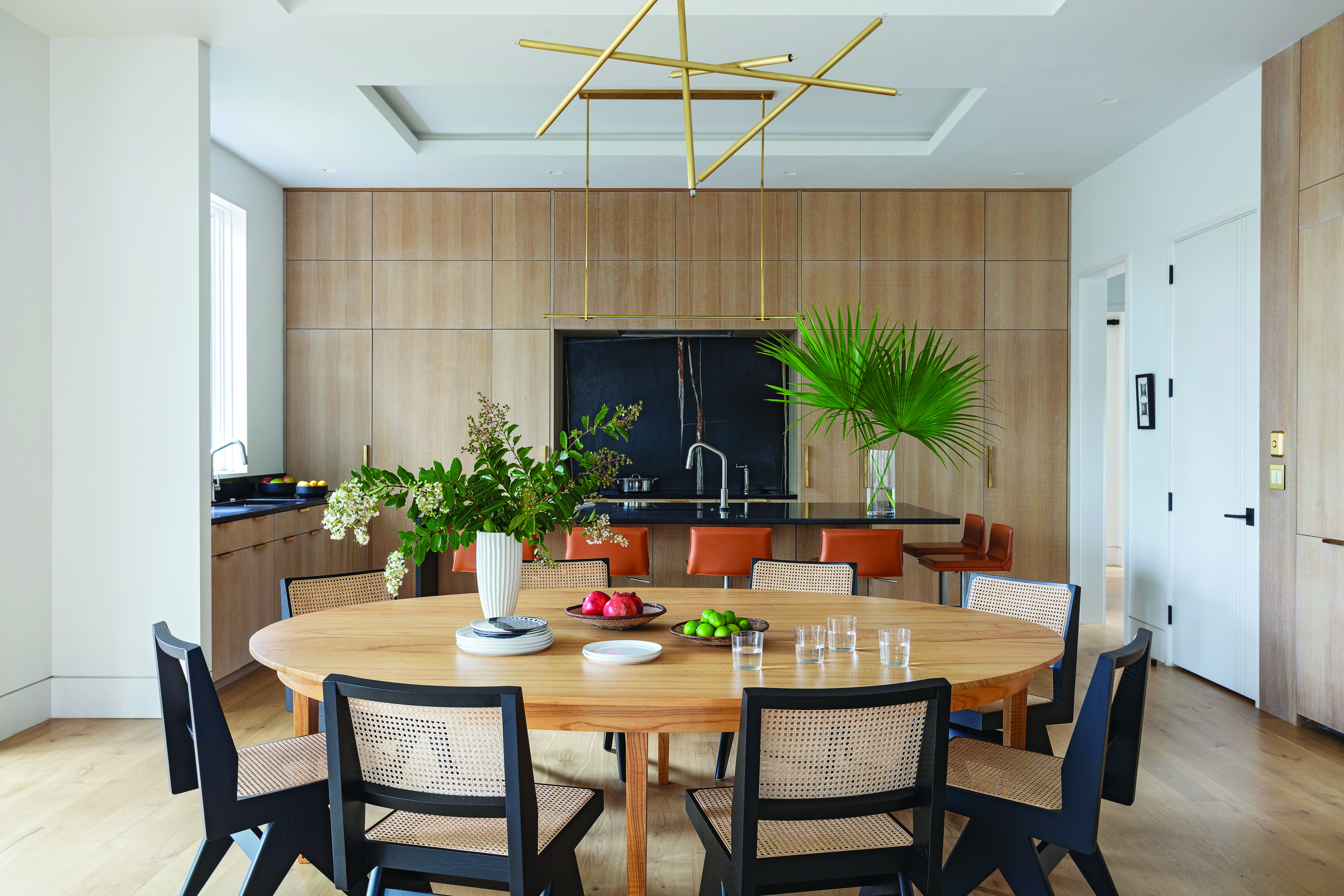 Modern Minimalism: The open dining/kitchen space is centered on the oval table the couple inherited from the prior owners of their midcentury modern home in Columbia. The table is accented by a custom Cam Crockford “pick-up stick” brass light fixture and black wicker chairs, which pull from the striking slab of Nero Dorato marble in the backsplash and quartzite countertops. An outdoor dining space can join the room thanks to accordion doors and retractable screens.