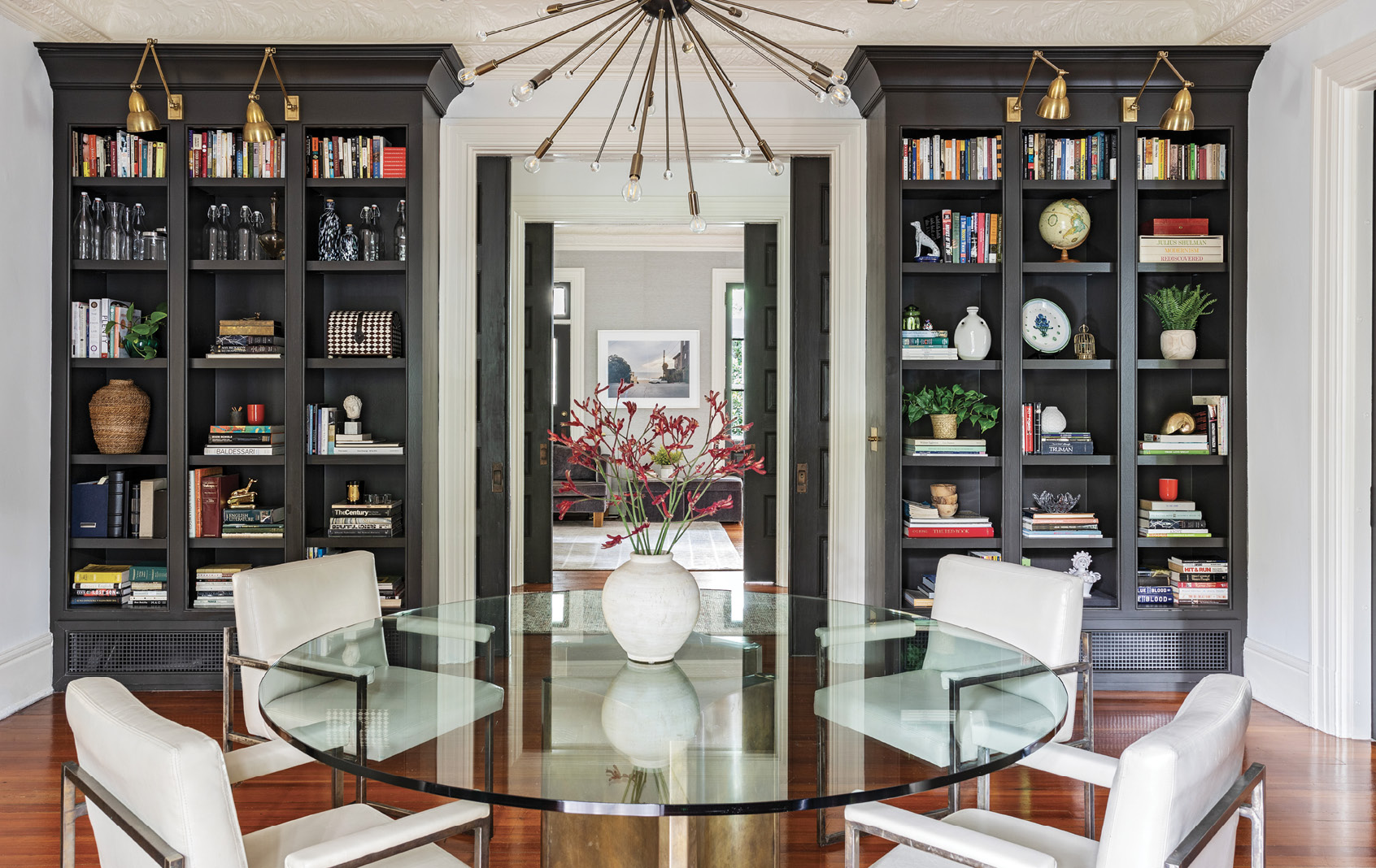 A large doorway leads to the dining room, where original built-in bookshelves, housing such gems as carved porcelain vases by local potter Maria White, frame a glass dining table atop a pair of brass Mastercraft table bases.