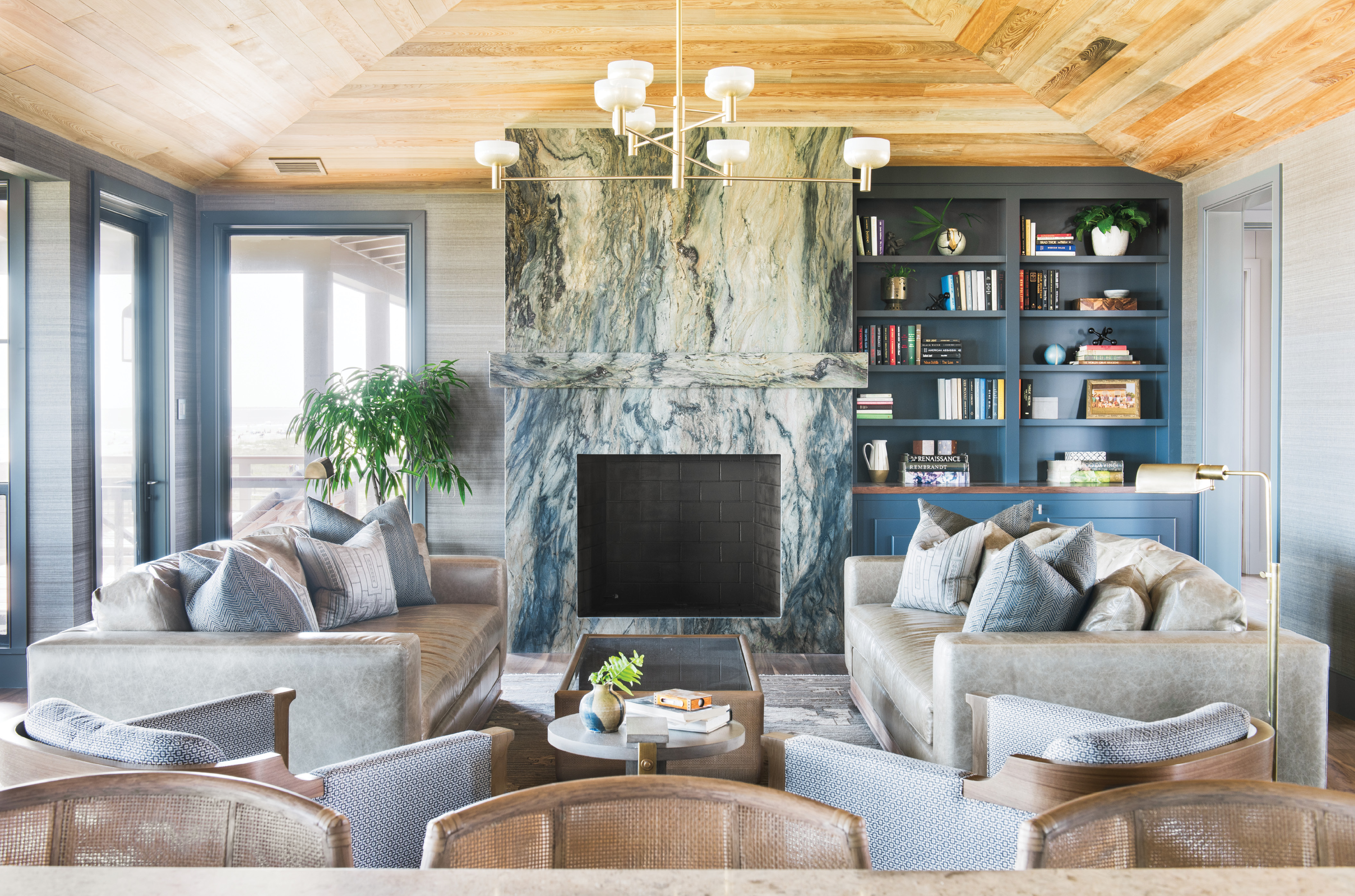 With the Tide: Situated on the top floor, the main living space is dominated by a fireplace set in “Blue Tides” granite from AGM Imports. Horsehair grasscloth wallpaper in “Clydesdale” by Phillip Jeffries continues the textural feel evoked by the stone’s veining. Leather sofas from Verellen and Osborne &amp; Little fabric-clad club chairs by Holly Hunt lend warmth and comfort to the scene, illuminated by Cartwright New York’s hand-brushed brass “Otto Luce” chandelier.