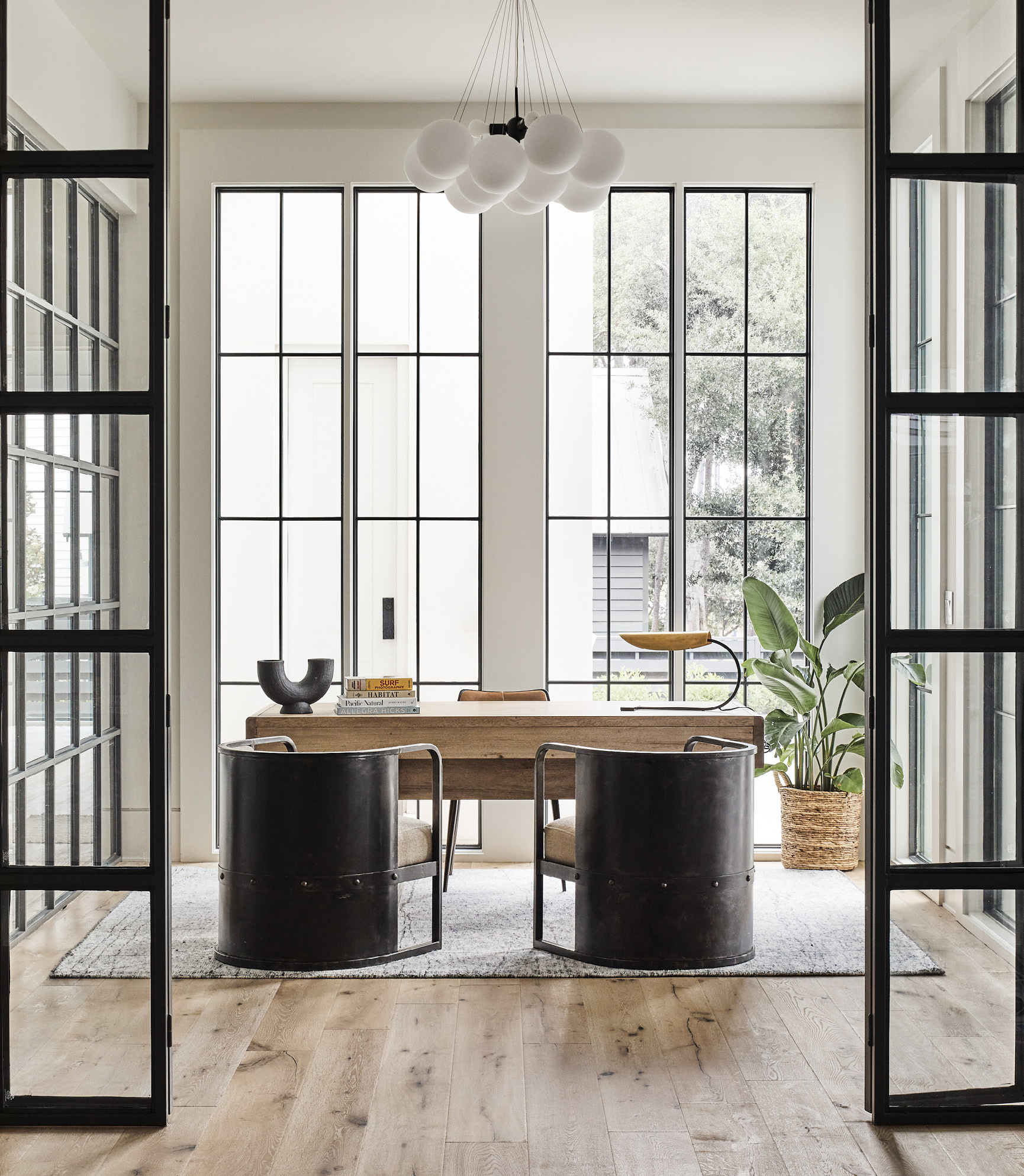Iron barrel-back chairs and a bulbous chandelier provide balance in the rectilinear iron and glass home office, where a washed walnut desk from Noir adds warmth.