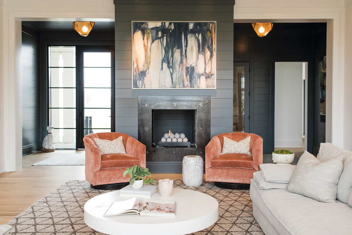The moody hues of the foyer—shiplap painted in Farrow &amp; Ball “Down Pipe”—continue into the living room around the dramatic fireplace. Crushed velvet chairs and an abstract painting by Jill Pumpelly add depth to the otherwise neutral palette.