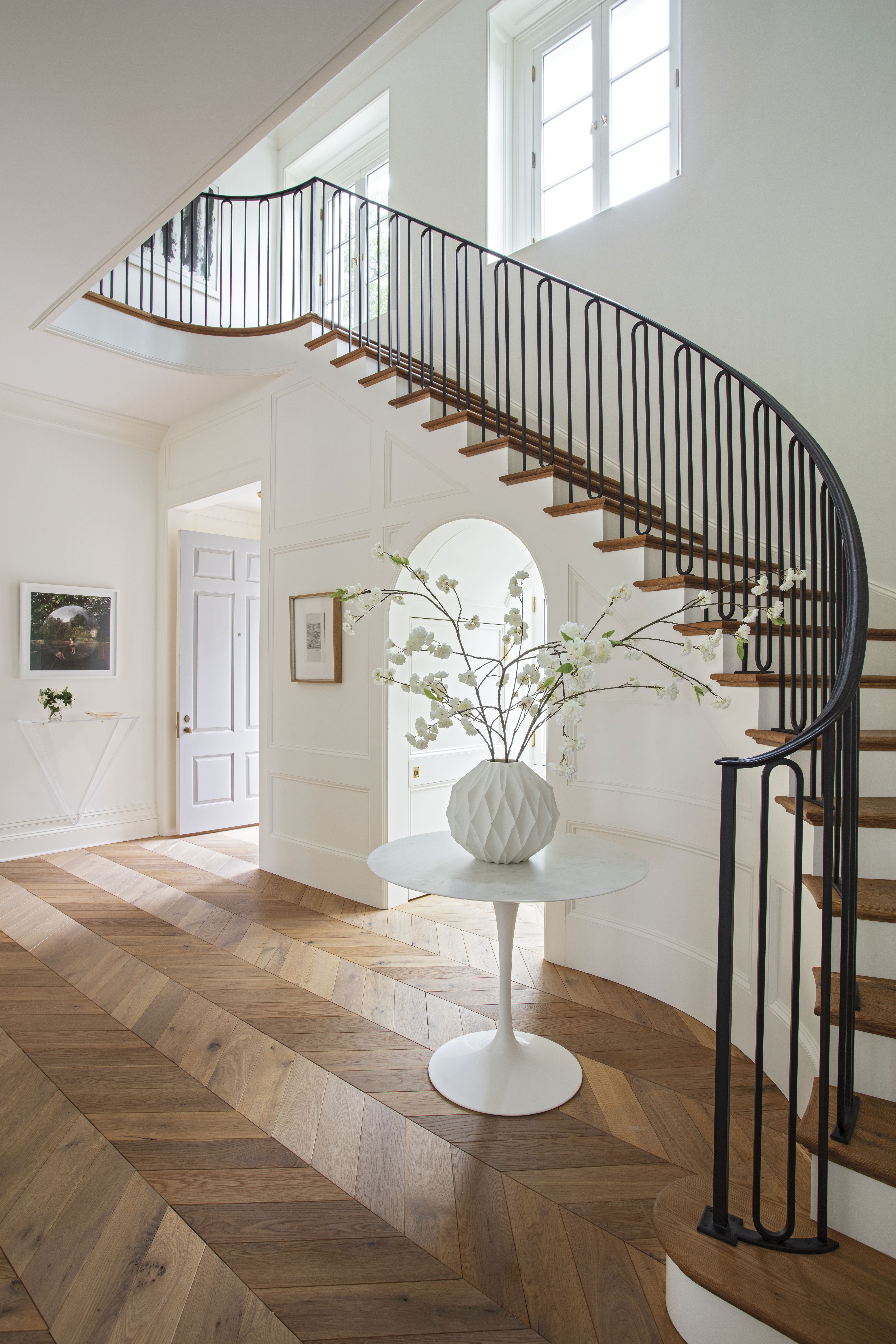 The showpiece entryway, dominated by a dramatic sweeping staircase, is flooded with natural light through a playful arched alcove and accented by white oak chevron flooring.