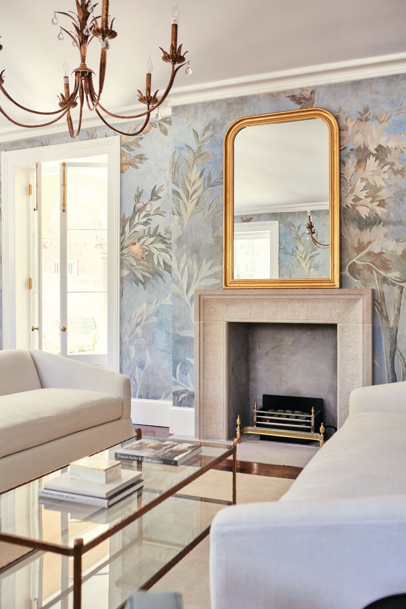 The formal drawing room, enveloped in the whimsical French Market Collection floral wallpaper, “De Soto Forrest,” balances classic and modern. Simple white sofas contrast with the dark heart of pine floors and antiques, and the Louis Philippe mirror above the carved limestone fireplace surround, antique brass coffee table, and elaborate Visual Comfort chandelier add subtle grandeur.