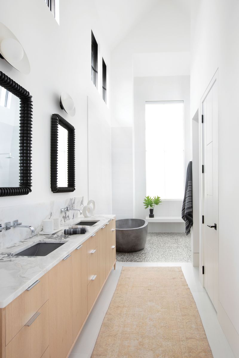 Natural light pours into the bathroom through a large window in the shower (with remote control shades). The warm wood of the vanity is contrasted by black-framed mirrors and marble countertops.