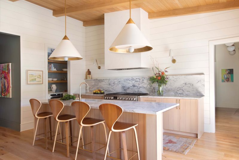 COLOR HER HAPPY:  The warm white kitchen, designed by Corin Havens, features shiplap walls, paneled white oak cabinets, marble countertops, and a custom plaster hood by Niki Miller of Charleston Fine Finishes. The space contrasts beautifully with the dark walls of the foyer, where herringbone floors and an artful grouping of rattan light fixtures add a natural element.