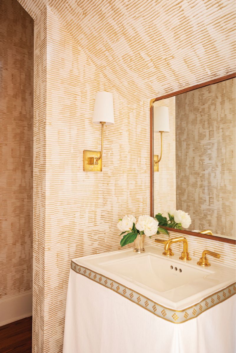A textured gold wallpaper from Romo in the nearby powder room adds pizazz, with its metallic tones picked up in the “Venini” gold sconce from Circa Lighting, the faucet, and the custom white linen sink skirt with a Samuel &amp; Sons trim.