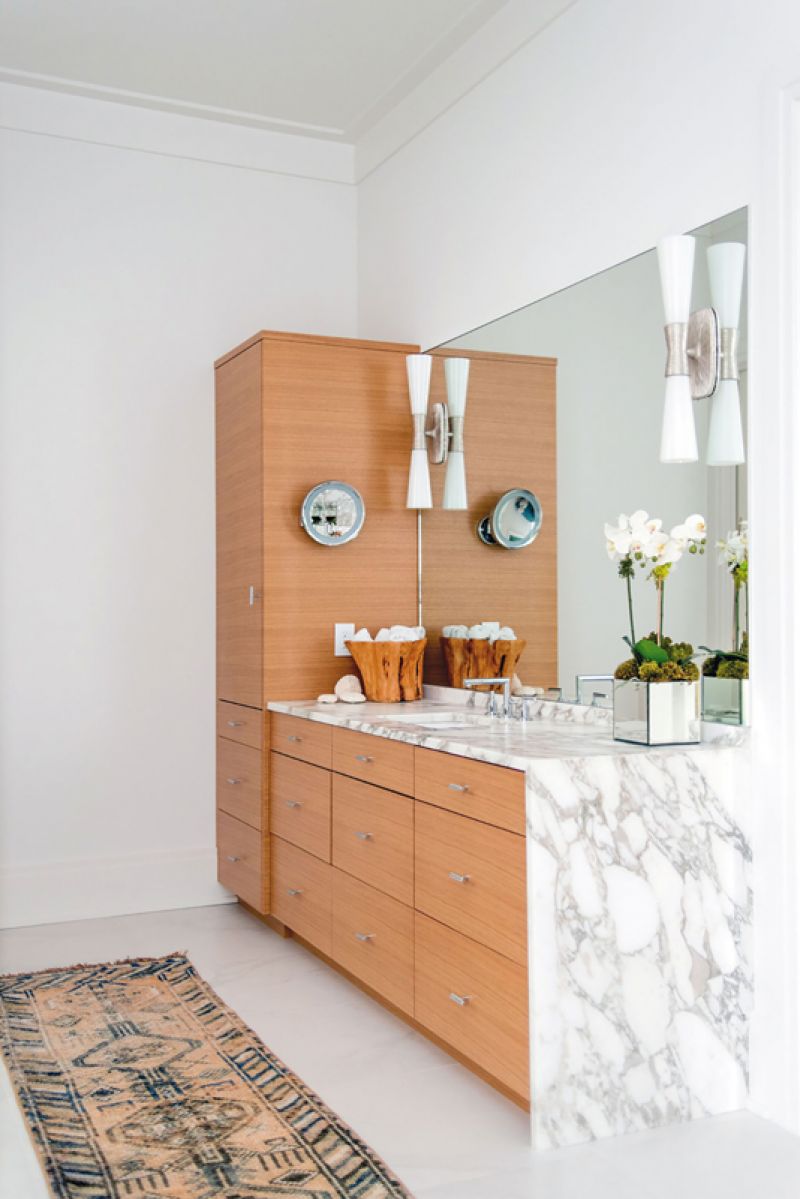 White oak cabinetry and a vintage runner warm the marble-clad bath, while sconces from Circa Lighting and an angular, custom-designed bench (over which a coastal scene snapped by Gray Malin hangs) add modern style in spades.
