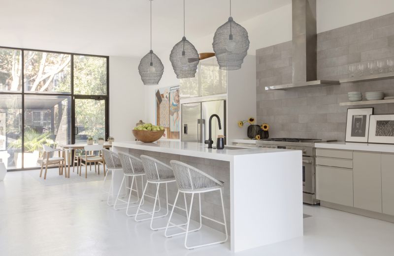 WHITE SPACE: Mixing textures in white, black, and beige, the kitchen is Baldwin’s quiet space. “With the black exterior and all white interior, I was drawn to a middle ground, and that manifested in a neutral-toned kitchen,” she says. “It allows you to have that burst of color as an offset.” Wire pendant lights from Denmark tie in to the industrial feel of the retail-style doors and windows; Teak Warehouse stools complete the look.