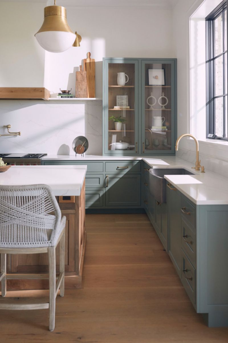 In the open kitchen, Water Street Brass hardware and durable quartz countertops complement the pale green cabinetry. A single glass-paned cabinet helps create symmetry with the plaster hood and the fireplace at the opposite end of the room.