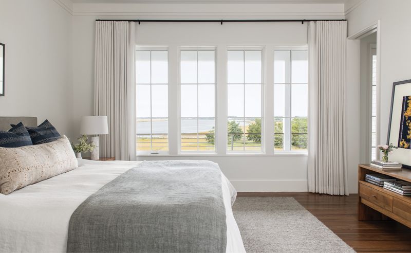 Marsh Mellow: Upstairs, the primary bedroom also takes advantage of the view with a similarly mellow color palette that accentuates the hues of the marsh beyond. White linen drapes, a soft gray rug from Armadillo Home, and custom linen bedding create a calm space.